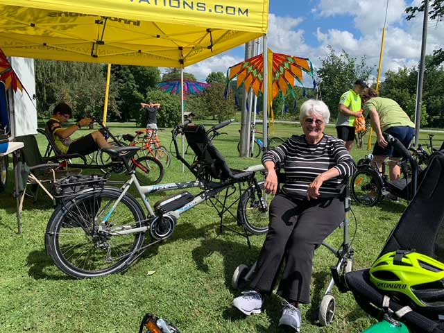 A smiling woman sits in a wheelchair in front of a Hase Pino tandem bike at the RAD-Innovations tent at the Cycle 4 CMT event